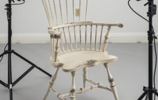 A vintage-inspired Painted Historical New England Comb Back Windsor Armchair featuring carved ears, knuckles, and a shaped seat. The legs are expertly mortised through the seat and wedged for durability. Finished in a soft linen paint, this armchair brings farmhouse charm and timeless elegance to any room. Perfect for adding a touch of historical craftsmanship to your home decor.